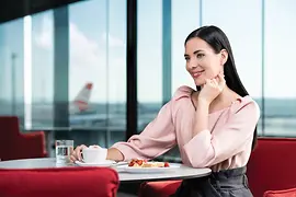 Mujer de cabello oscuro en el lounge de Austrian Business en el Aeropuerto de Viena que espera a embarcar mientras disfruta de una taza de Melange y una porción de tarta.